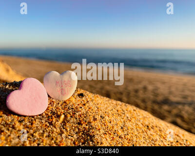 Deux bonbons en forme de coeur de Saint-Valentin sur une plage ensoleillée. L'une des bonbons a des mots à manquer sur elle. Banque D'Images