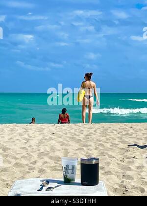 Boissons au premier plan sur le sable, une femme dans un bikini noir portant un sac à main jaune se tient près de l'eau turquoise Miami South Beach, Floride, États-Unis Banque D'Images