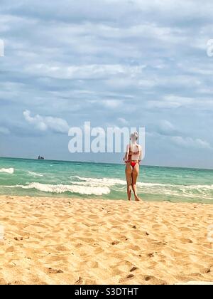 Une femme blonde dans un bikini rouge marche sur le sable vers l'eau turquoise de l'océan avec un bateau de croisière à l'horizon à Miami South Beach, Floride, Banque D'Images