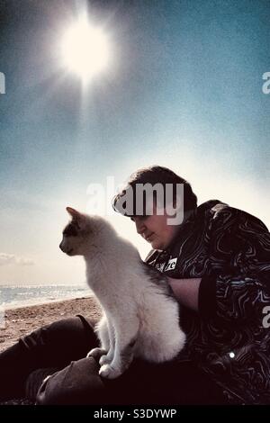 Un adolescent assis sur une plage avec un chat qui s'ébouillante sur les genoux Banque D'Images