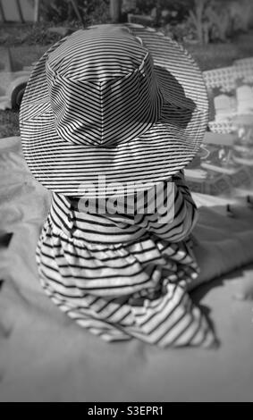 Une petite fille avec une robe strippy et un chapeau joua sur un tapis dans le jardin avec son dos à la caméra Banque D'Images