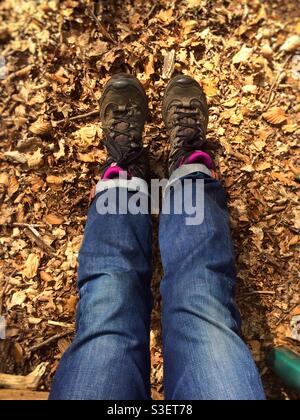 Les jambes d’une femme sont étirées devant elle alors qu’elle est assise sur le sol forestier parsemé de feuilles. Elle porte des jeans bleus et des bottes de randonnée. Banque D'Images