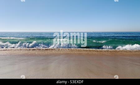 La vague de l'océan se brise sur une plage de sable sous un ciel bleu clair. Banque D'Images