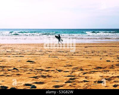 Un surfeur de ligne transportant une planche de surf le long d'une plage de sable n Cornwall avec l'océan derrière Banque D'Images