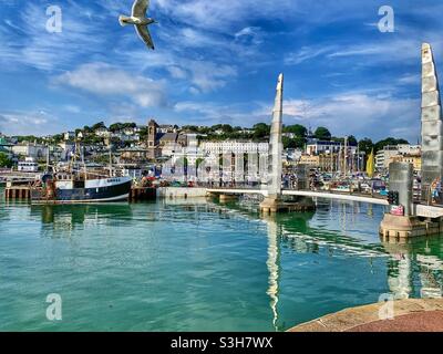 L'été au port de Torquay. Un mouette survole la ligne du ciel de la ville par une journée ensoleillée. Eau turquoise. Banque D'Images