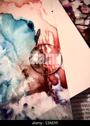 Une partie de peinture abstraite à l'encre d'alcool avec des couleurs rouge, aqua, rose, marron et pourpre sur du papier journal et du papier blanc sur une surface plate avec une cachochon de verre rond sur une partie de l'encre d'alcool. Banque D'Images