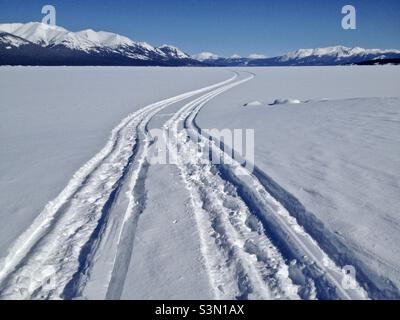 Deux pistes de motoneige sur le lac Atlin recouvert de neige, avec ciel bleu azur et montagnes en arrière-plan. Banque D'Images