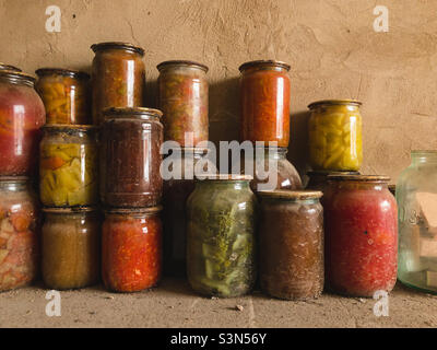 Canned vegetables, fruits and salads in glass jars under an iron lid stand on a dusty floor in a cellar in a garage. Stock Photo