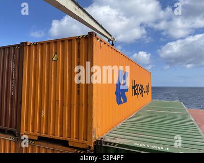 Conteneur orange de la société Hapag Lloyd, Hambourg, Allemagne, chargé et écrasé sur un conteneur avec des grues. Le navire est en cours de route par temps ensoleillé et calme, avec un ciel bleu et des nuages blancs. Banque D'Images