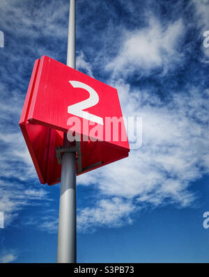 Chiffre blanc numéro deux sur un signe triangulaire rouge vif suspendu sur un poteau haut avec contre un ciel bleu brillant. Banque D'Images