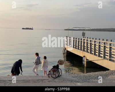 Famille avec fauteuil roulant joue sur la plage de galets nicht en bois petit quai ou jetée pendant le coucher du soleil dans le port de Koper, Slovénie. À l'horizon se trouve le littoral et le navire marchand amarré. Banque D'Images