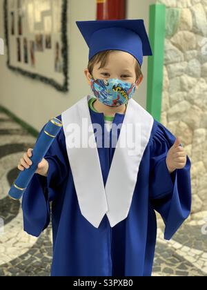 Un jeune garçon heureux en casquette bleue et robe portant un masque lors de sa remise des diplômes de maternelle pendant la pandémie de covid. São Paulo, Brésil. Banque D'Images