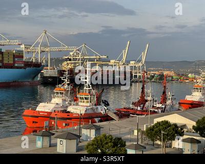 Deux remorqueurs à coques orange amarrés dans le port de Koper, en Slovénie. En arrière-plan se trouvent deux conteneurs lors de l'opération de chargement par des grues portiques sous ciel bleu. Banque D'Images