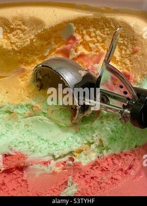 Une cuillère à crème glacée dans un gallon de crème glacée Rainbow Sherbert prêt à commencer à remplir tous les bols et les cônes des amateurs de crème glacée. Banque D'Images