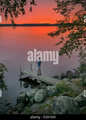 Un garçon prenant une photo du coucher de soleil sans fin pendant la période de soleil de minuit de l'été dans un lac d'été près du cercle arctique en Finlande Banque D'Images