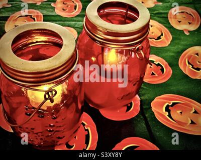 Une paire de bougies orange en pot de maçon donne un scintillement de lumière assis sur une table recouverte d'une nappe en forme de citrouille d'Halloween saisonnière. Banque D'Images