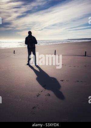 Homme rétroéclairé debout sur une plage à marée basse Banque D'Images