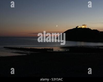 Ciel nocturne avec un croissant de lune visible et le château de Criccieth en arrière-plan, éclairé au sommet d'une colline surplombant la mer. Banque D'Images