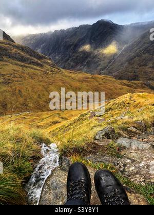 Vue sur les pieds et les bottes Walkers avec des puits de lumière comme les nuages commencent à se briser sur le chemin de Munro Buachaville Etive Beag, avec une vue vers la crête de Aonach Eagach crête, Glencoe, Écosse Banque D'Images