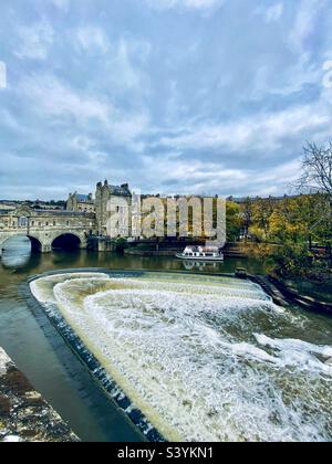 Pont Pulteney sur la rivière Avon à Bath, Angleterre, Royaume-Uni. Le pont est l'un des quatre seuls ponts dans le monde qui a des magasins sur toute sa travée des deux côtés. Déversoir en fer à cheval au premier plan. Banque D'Images