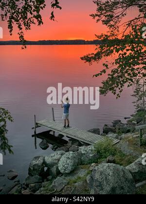 Un adolescent prend une photo avec son téléphone portable de soleil de minuit et un ciel rouge de sang au-dessus d'un quai de lac dans une maison d'été dans la région de Kajaani en Finlande Banque D'Images