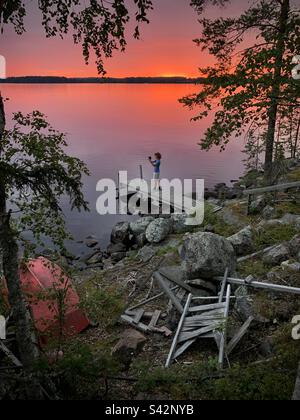 Un adolescent prend une photo avec son téléphone portable de soleil de minuit et un ciel rouge de sang au-dessus d'un quai de lac dans une maison d'été dans la région de Kajaani en Finlande Banque D'Images
