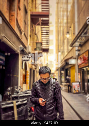 Jeune homme asiatique utilisant un téléphone portable tout en se tenant à côté de vélos de livraison de nourriture dans une allée contre des bâtiments dans la ville de Melbourne, Australie. Banque D'Images