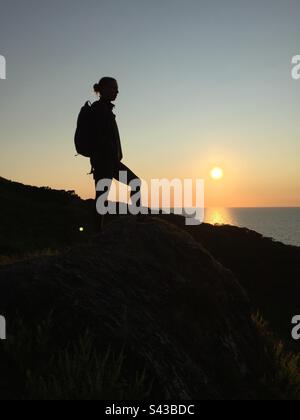 Une femme en silhouette au coucher du soleil se promenant au-dessus de la mer d'Irlande sur une section spectaculaire du célèbre Ceredigion Coast Path près de Llangrannog dans l'ouest du pays de Galles Banque D'Images