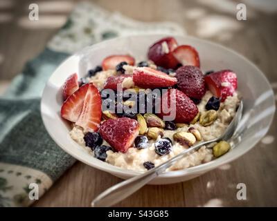 Flocons d'avoine fraîchement cuits avec fraises en tranches, bleuets séchés et pistaches dans un bol blanc avec serviette verte sur une table en bois. Banque D'Images