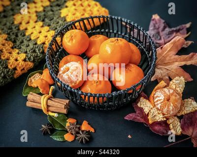 Mandarines pelées et entières dans un panier noir entouré d'épices, d'herbes, de feuilles rouges séchées et de lainages crotchés sur fond noir. Thème de l'automne. Encore la vie. Fruits. Banque D'Images