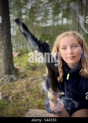 Portrait d'une adolescente de 11 ans à un feu de camp dans la nature en profitant de la vie en plein air avec un camping en bâton brûlant à la maison d'été dans la région de Kajaani en Finlande pendant un été arctique Banque D'Images