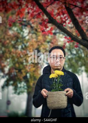 Portrait d'un jeune homme asiatique en lunettes portant une plante à fleurs en pot de chrysanthème jaune dans un panier lors d'un jour d'automne brumeux sous des arbres avec feuillage d'automne. Banque D'Images