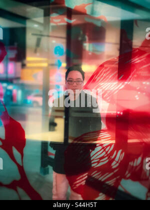Différents niveaux de réalité. Portrait d'un jeune homme asiatique debout derrière un écran en verre avec reflets et éclairage au néon sur la rue de la ville la nuit. Surréaliste. Rêveur. Banque D'Images