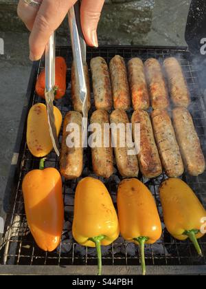 Cuisson des saucisses et des poivrons sur le barbecue extérieur, gros plan avec la main de la femme et les pinces dans le cadre Banque D'Images