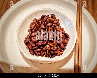 Vue en grand angle des graines de citrouille assaisonnées dans un bol sur l'assiette avec des baguettes sur la table. Banque D'Images
