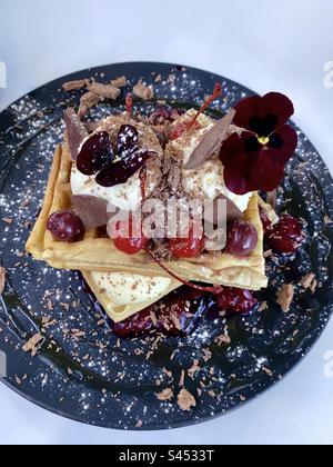 Gaufres Forêt noire : deux gaufres avec crème glacée, cerises et chocolat sur une assiette noire. Banque D'Images