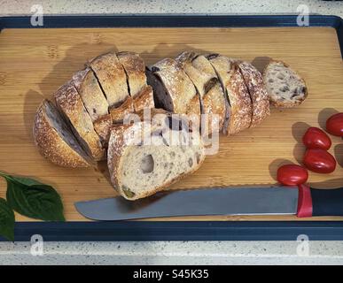 Vue à angle élevé de pain au levain d'olive tranché et croustillant avec tomates cerises, basilic et couteau à pain sur une planche à découper en bois sur le comptoir. Banque D'Images