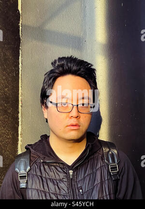 Portrait de jeune homme asiatique en lunettes contre un mur. Soins de la vue, coiffure. Lumière du soleil et ombres. Banque D'Images