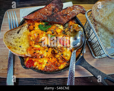 Vue à grand angle des œufs cuits au four avec des haricots, des tomates et du chorizo dans une poêle avec du pain au levain sur une planche de bois sur la table. Banque D'Images