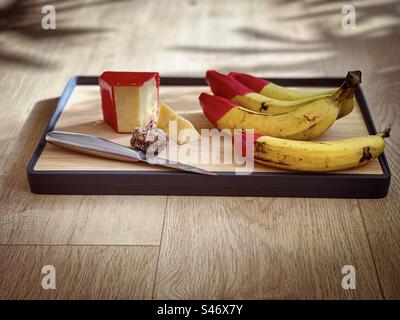 Fromage cheddar vieilli avec revêtement de cire rouge, une figue séchée, des bananes bio, à pointe de cire rouge et un couteau sur une planche à découper en bois sur la table avec espace de copie. Lumière du soleil et ombres. Banque D'Images