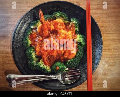 Directement au-dessus de la vue de crevettes sautées dans la sauce Chili épicée avec du brocoli dans une assiette noire avec des baguettes rouges et fourchette et cuillère sur la table en bois. Cuisine chinoise. Cuisine. Gourmet. Banque D'Images