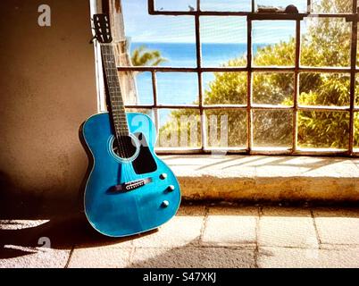 Guitare bleue appuyée contre un cadre de fenêtre cassé donnant sur une mer bleue Banque D'Images