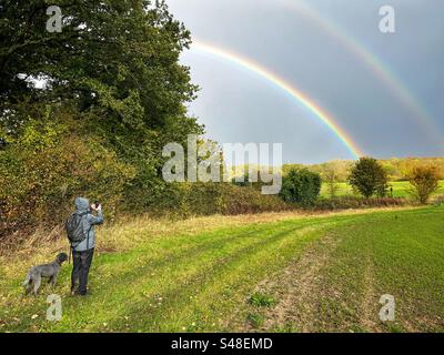 Une femme s'arrête pour prendre une photo d'un arc-en-ciel tout en promenant son chien labradoodle miniature le long d'un sentier dans le Hampshire rural en Angleterre. Banque D'Images