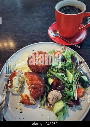 Vue à angle élevé des galettes de saumon avec sauce hollandaise et salade fraîche sur assiette avec couverts et café noir dans une tasse rouge et soucoupe sur table. Banque D'Images