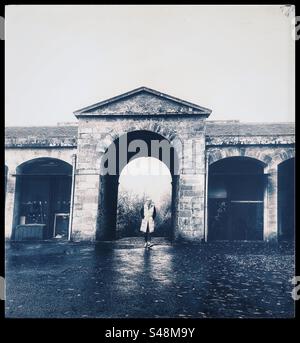 Photo en noir et blanc d'une femme marchant à travers une arche de brique sur une ancienne ferme. Banque D'Images