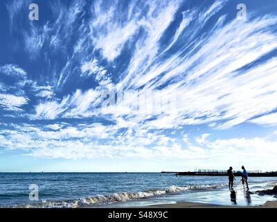 Des nuages plumeux étonnants traînant dans un ciel bleu pendant l'été, avec deux personnes face à face sur la plage. Banque D'Images
