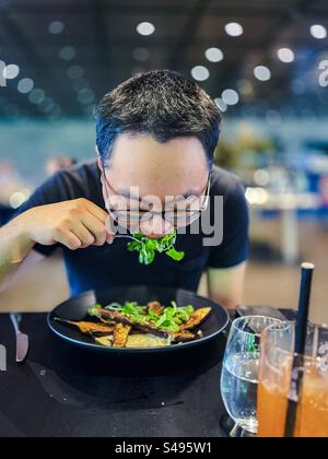 Un homme en lunettes appréciant un repas de salades fraîches et des aubergines rôties marinées au miso à table dans le restaurant. Manger sainement. Repas végétarien. Concentrez-vous sur le premier plan. Banque D'Images