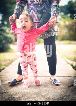 Les premiers pas de bébé à l’extérieur. Bébé marchant avec excitation étant aidé par un adulte Banque D'Images
