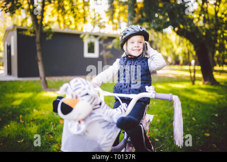 Enfant drôle Caucasian girl blonde dans un casque de vélo à proximité d'un vélo violet avec un panier à l'extérieur du parc, sur une pelouse verte panier d'herbe à la maison. Joue u Banque D'Images