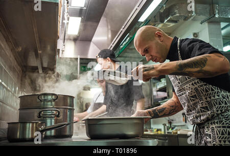 Une cuisine de restaurant. Chef professionnel en tablier, avec des tatouages sur ses bras est le tamisage de la farine en cours de cuisson avec ses deux assistants. Cuisson Banque D'Images